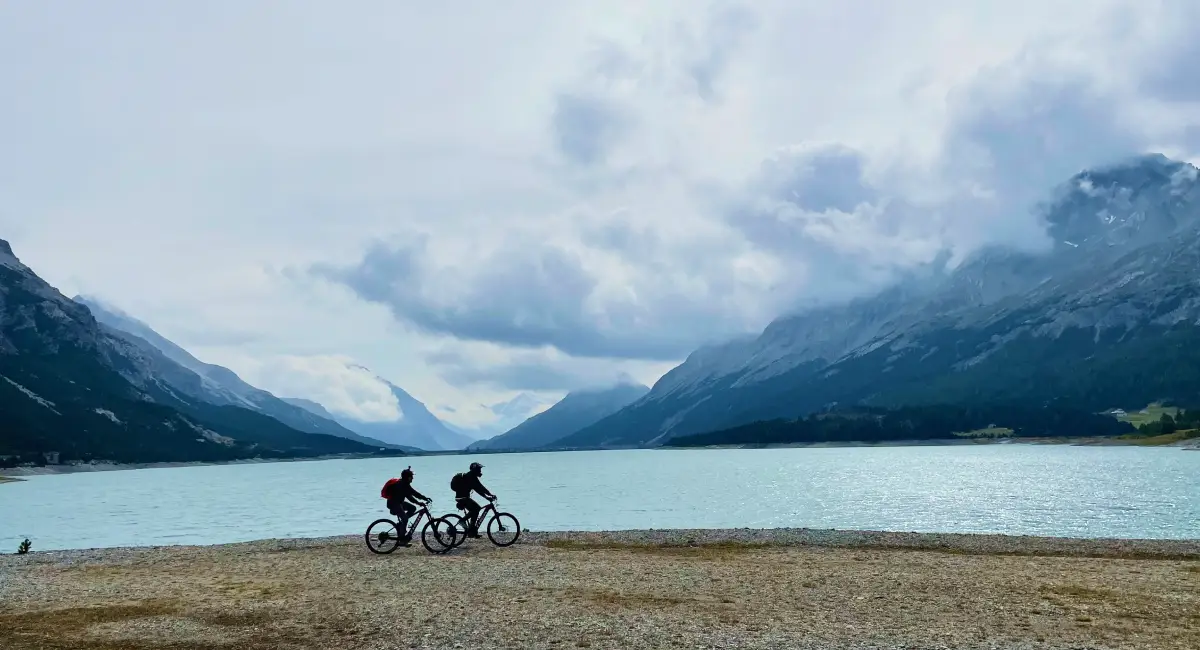 Two cyclists riding along the turquoise waters of Cancano Lake surrounded by dramatic peaks