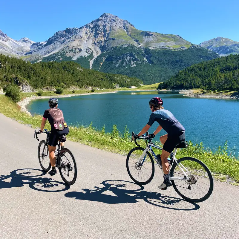 Two cyclists riding along Cancano Lake surrounded by alpine peaks and crystal-clear water