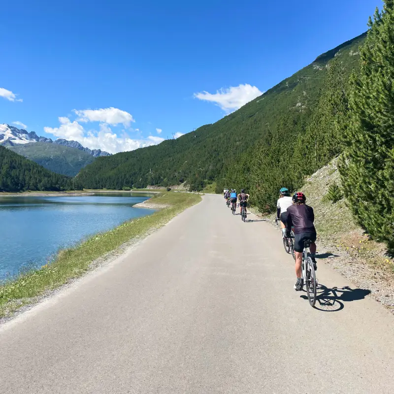 Cyclists riding along the turquoise Cancano Lakes near Bormio surrounded by alpine peaks during the Epic Alps bike tour