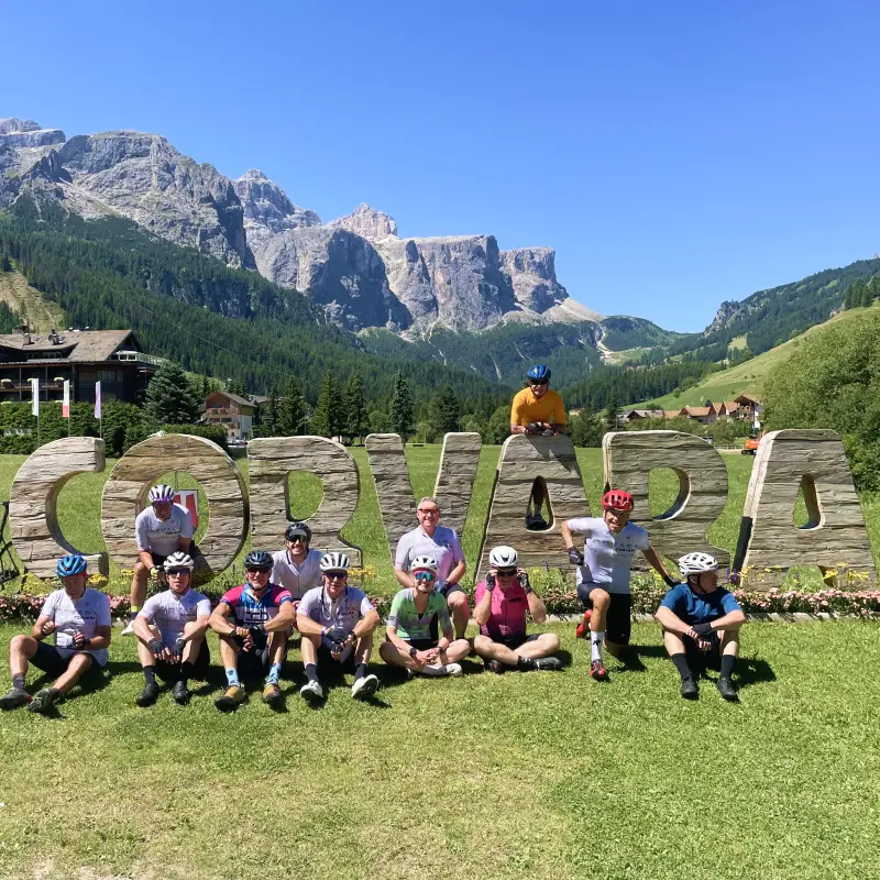 Group of cyclists posing by the Corvara sign with the Sella massif in the background