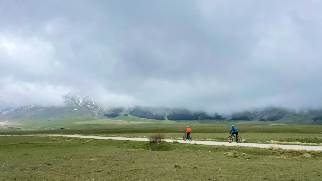 Two cyclists riding through the misty plains of Campo Imperatore under dramatic clouds