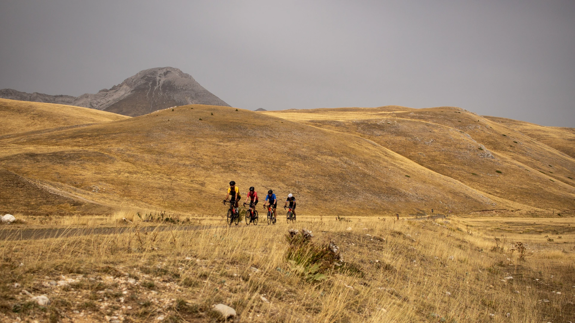Cyclists riding in a line across the golden hills of Campo Imperatore under dramatic skies