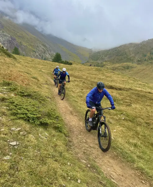 Group of cyclists riding e-MTBs down a scenic mountain trail surrounded by meadows and clouds
