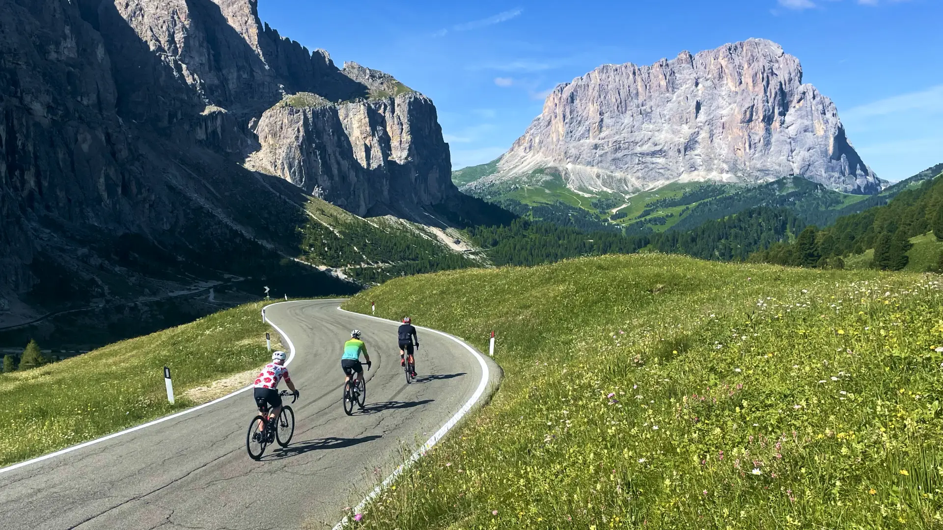 Cyclists riding along a winding mountain road surrounded by towering Dolomite peaks and alpine meadows on the Epic Dolomites Bike Tour