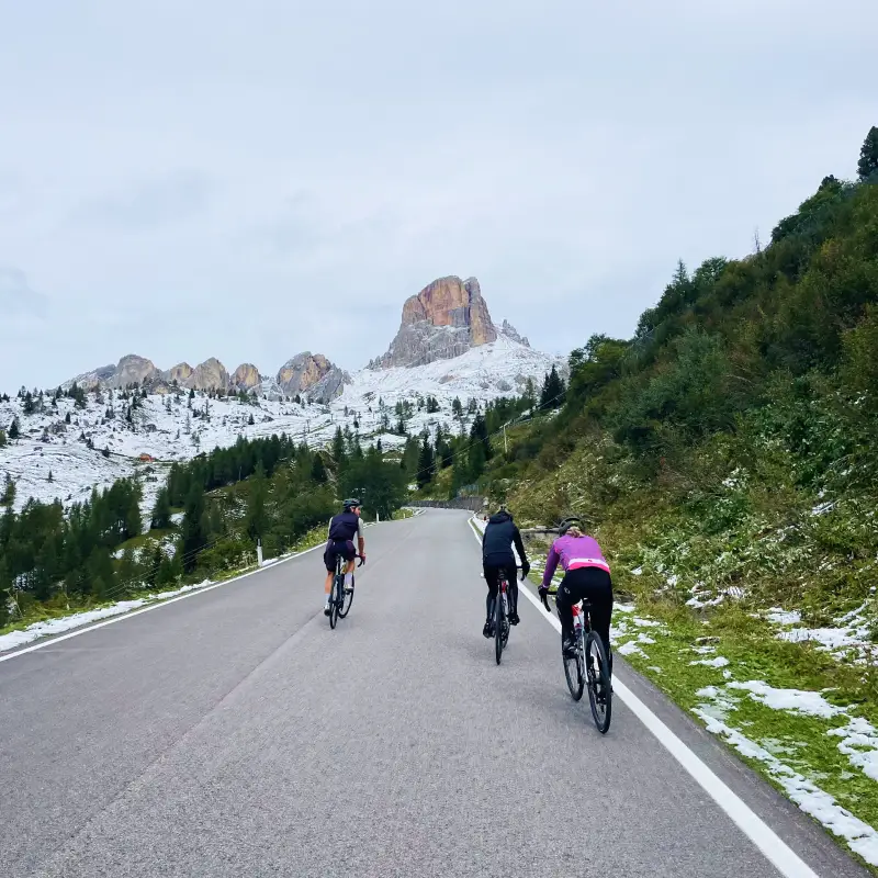 Three cyclists climbing the snowy Passo Giau with Cinque Torri in the background