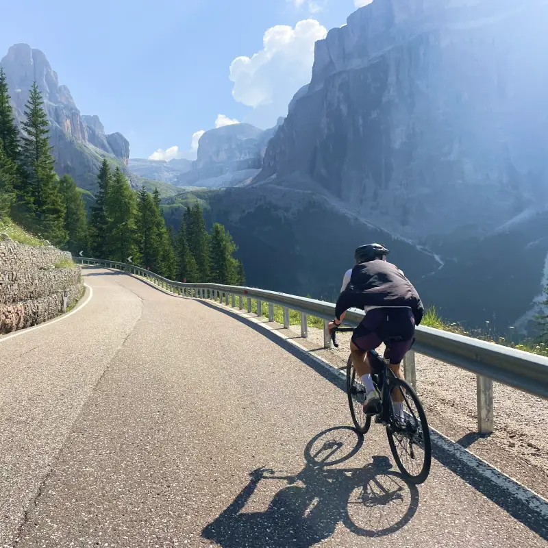 Cyclist riding uphill toward the Sella Pass surrounded by dramatic Dolomite cliffs and pine forests