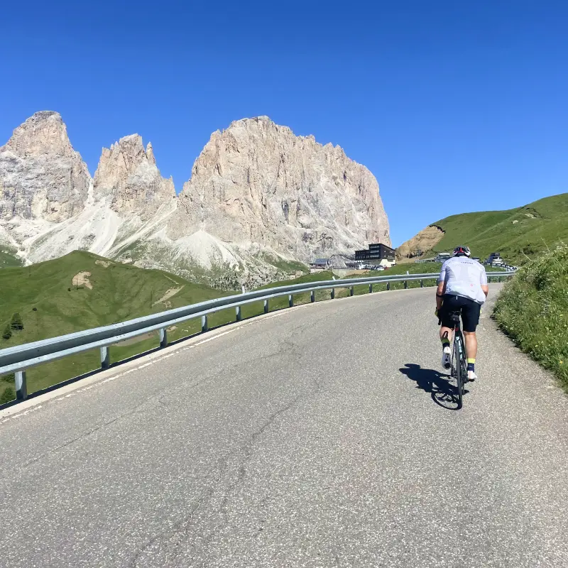 Cyclist climbing toward Passo Sella surrounded by the jagged Dolomites peaks under a bright blue sky